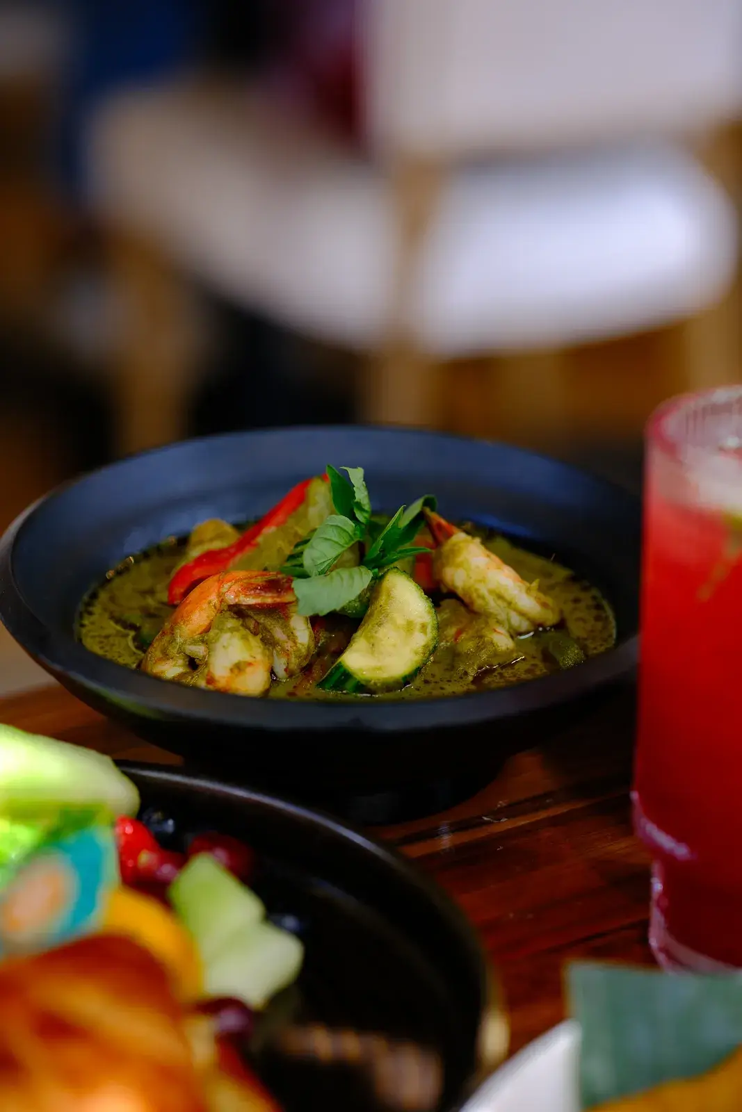 A brunch table crowded with plates, a green curry, fried rice and a red iced drink