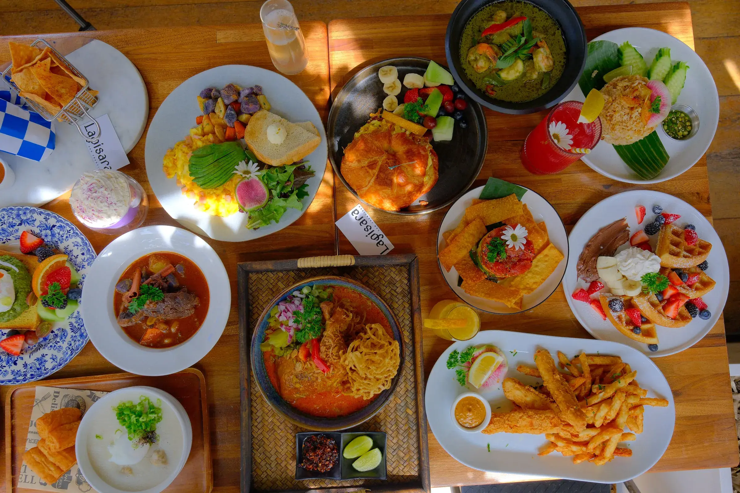 A table of brunch plates seen from above: rice bowls, noodles, fried chicken, eggs and iced drinks